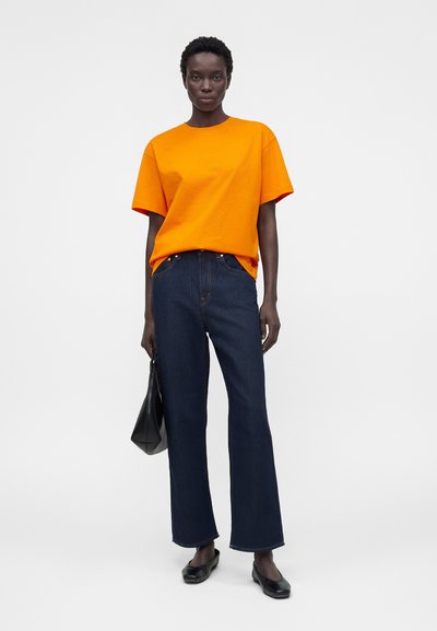 Personne portant une chemise à manches courtes orange vif, un jean droit bleu foncé, des chaussures noires, tenant un sac à main noir devant un fond uni.