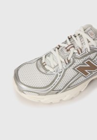 Harmaa ja beige New Balance urheilujalkine, jossa on verkkokangasosa, ruskea logo ja valkoiset nauhat, esitetty lähikuvassa valkoista taustaa vasten.