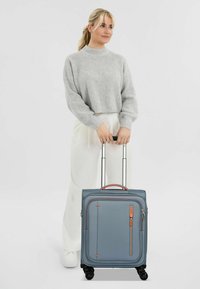 Valigia con ruote in tessuto azzurro chiaro con dettagli arancioni, dotata di due manici telescopici e una tasca con zip frontale. Design liscio e strutturato.