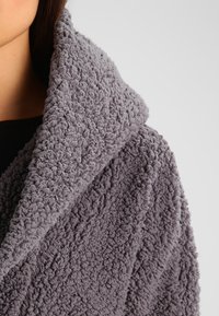 Accappatoio grigio, in tessuto morbido e con texture, dotato di un ampio colletto, caratterizzato da un aspetto soffice e accogliente e una superficie leggermente stropicciata.