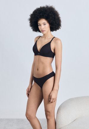 Kvinde med afro-frisure iført sort bh og undertøj, stående ved siden af en hvid tekstureret lænestol mod en ensfarvet baggrund.