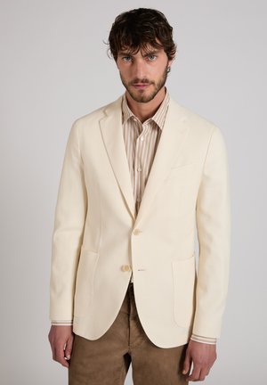 Mann mit dunklem Haar und Bart trägt ein cremefarbenes Blazer über einem beigen gestreiften Hemd und braunen Hosen vor einem schlichten hellen Hintergrund.