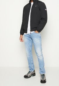 Giacca isolante nera con colletto alto, abbinata a jeans strappati blu chiaro e sneakers grigie texture con accenti colorati.