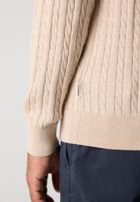 Μπεζ πουλόβερ με πλέξη καλωδίου, με μανίκια και τελείω�μα πάνω από σκούρα μπλε παντελόνια, εμφανίζοντας μια μικρή λευκή ετικέτα μάρκας στη ραφή του πουλόβερ.