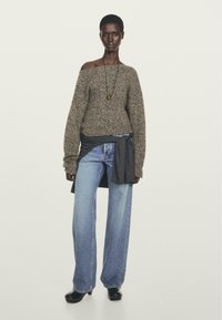 Donna che indossa un maglione melange con le spalle scoperte, jeans azzurri a gamba larga, zoccoli neri, una lunga collana a pendente e una felpa scura legata in vita.