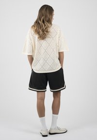 Personne aux longs cheveux ondulés portant un haut blanc texturé à manches courtes, un short noir avec une bordure blanche, des chaussettes blanches et des mocassins blancs, debout, de dos.