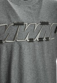 Camiseta gris con un destacado diseño bordado en un gris más claro, con tres líneas verticales de acentos de costura negra.