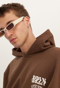 Jauns vīrietis, kas valkā nelielas taisnstūra formas baltas saulesbrilles, riņķa auskaru un brūnu kapuci ar baltu grafisku tekstu un pasaules ikonu.