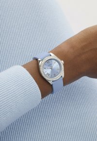 Relógio com bracelete de silicone azul claro, caixa prateada, mostrador redondo com marcadores de números romanos, adornado com cristais cintilantes.