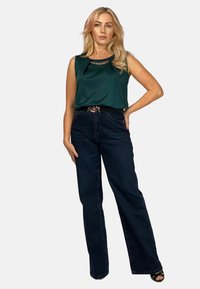 Jeans w ciemnym dżinsie w połączeniu z satynowym, turkusowym topem bez rękawów z łańcuszkowym detalem przy szyi. Modelka stoi w zrelaksowanej pozycji.