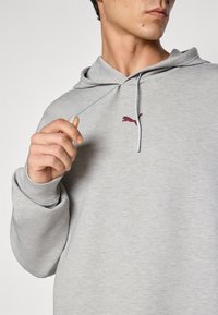 Mand iført en lysegrå hættetrøje med maroon Puma-logo, der holder og trækker lidt i en snor nær brystområdet.