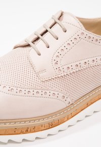 Chaussure brogue en cuir beige clair avec détails perforés, panneaux en tissu, fermeture à lacets et semelle superposée en liège et caoutchouc blanc.