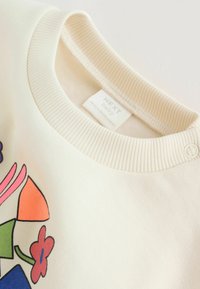 Sudadera para bebés color crema con cuello acanalado, que presenta formas geométricas coloridas y diseños florales. Etiqueta visible en el interior.