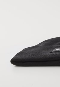 Zwarte gebreide beanie met een geribbelde textuur, omgeslagen rand en een klein merklabel aan de voorkant. Het materiaal lijkt zacht en flexibel.
