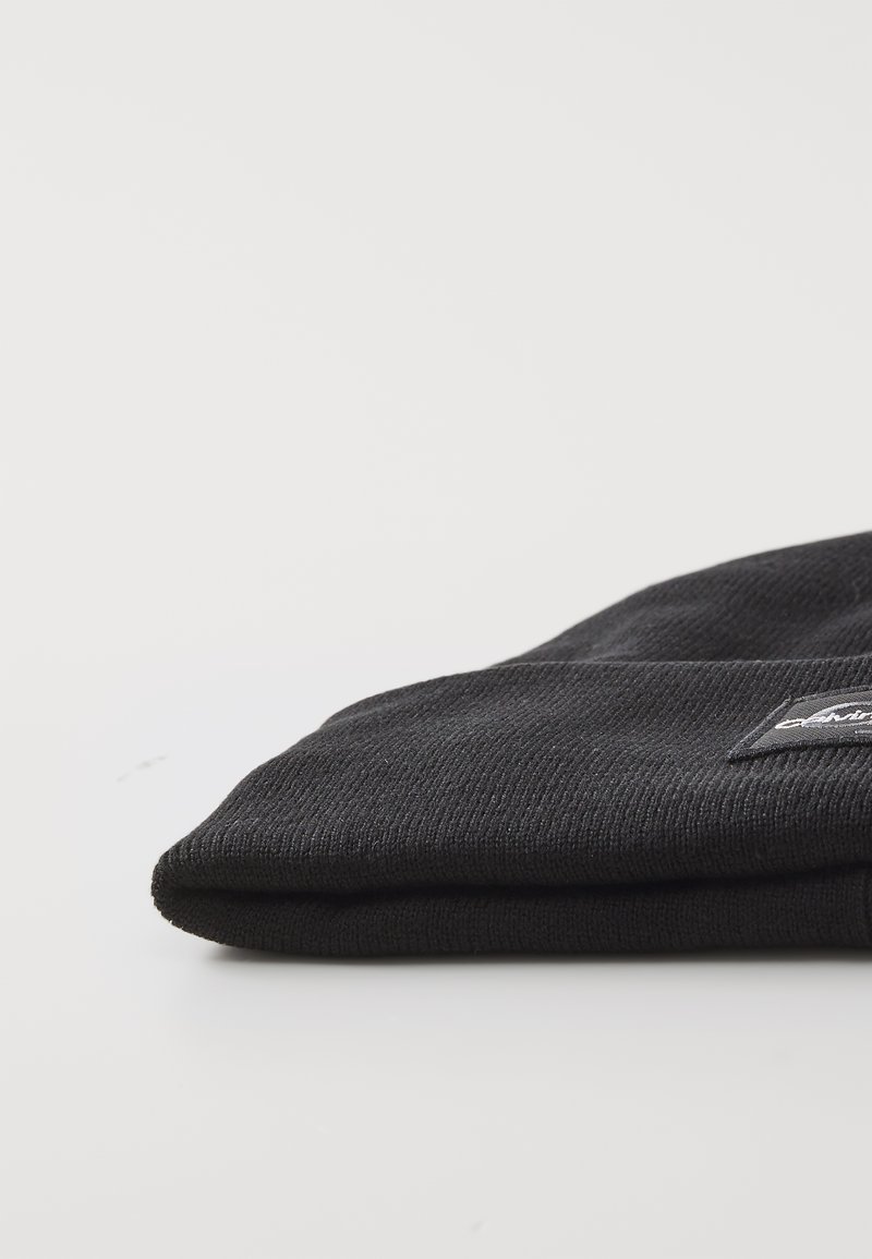 Czarna dziana czapka typu beanie z ryflowanym wzorem, złożonym brzegiem i małą łatką z logo z przodu. Materiał wydaje się miękki i elastyczny.