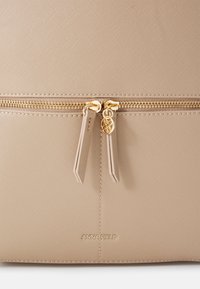 Borsa a tracolla sintetica beige con chiusura a zip dorata, superficie texturizzata e logo del marchio sul fondo. Gli accessori includono tiranti della zip coordinati.