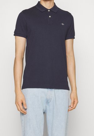 Homme portant un polo bleu marine ajusté à deux boutons et un jean bleu clair, debout devant un fond blanc uni.