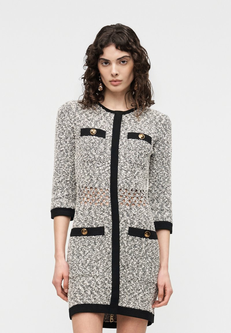 Woman with curly hair wearing a textured gray dress with black trim, gold buttons, and a patterned waist cutout, standing against a plain background.