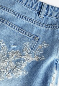 Vaate siniseen denim-kankaaseen, jossa on kukkakuvioista valkoista brodeerausta ja pieni repeämä, sekä vyötärö ja takatasku.