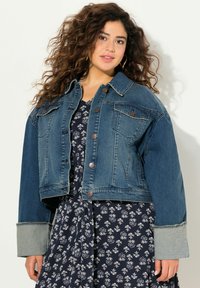 Giacca di jeans cropped blu scuro con bottoni, due tasche sul petto e polsini azzurri a contrasto. Indossata sopra un vestito floreale.