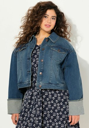Chaqueta de mezclilla oscura recortada con botones, dos bolsillos en el pecho y puños en un azul claro contrastante. Llevada sobre un vestido floral.