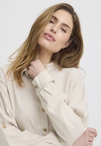 Jeune femme aux cheveux châtain clair portant une chemise beige boutonnée, touchant doucement le col avec une expression calme sur un fond uni.