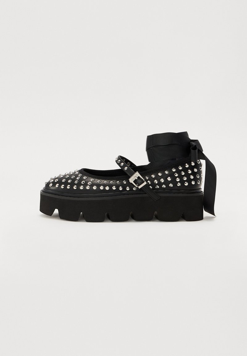 Black platform shoes with a rounded toe, embellished with metal studs, and an ankle strap tied with a satin ribbon. Textured rubber sole.