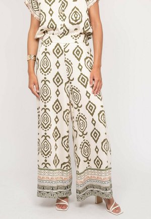 Pantalon à jambes larges avec des motifs géométriques vert olive et beige, associé à des talons blancs à brides et un haut assorti à motifs.