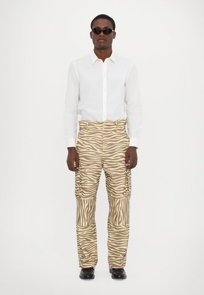 Beige en bruine zebra-gestreepte cargo broek met zakken; gecombineerd met een wit blouse-shirt en zwarte zonnebril. Eenvoudige achtergrond.