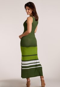 Žena sa úsmevom cez rameno, oblečená v zelených maxi šatách bez rukávov s horizontálnymi pruhmi pri okraji a hnedými klinovými sandálmi.