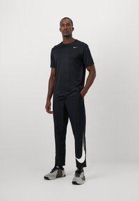 Sort atletisk t-shirt med rund hals, parret med sorte træningsbukser med et stort hvidt Nike-logo på siden og grå sneakers.