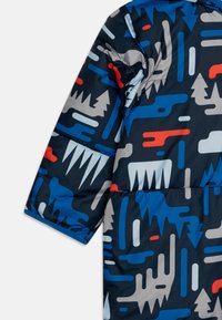 Veste matelassée avec un motif abstrait bleu, rouge et gris comportant des formes d'arbres et de vagues sur fond sombre. Tissu texturé, manches courtes.