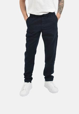 Uomo con pantaloni casual neri a vita elastica, scarpe da ginnastica bianche, mani nelle tasche e avambracci tatuati su sfondo semplice.