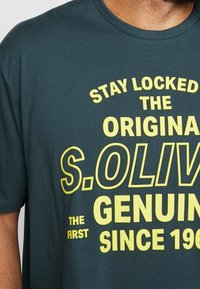 Donkerteal katoenen T-shirt met gedurfde gele tekst: "STAY LOCKED THE ORIGINAL S.OLIV." en "GENUINE THE FIRST SINCE 1980."