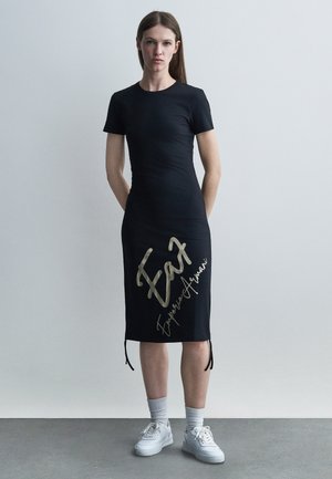 Vrouw staand, draagt een zwarte midi-jurk met korte mouwen en de tekst "EA7 Emporio Armani", gecombineerd met witte sneakers en sokken.