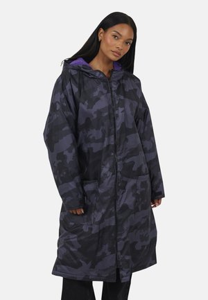 Donna che indossa un lungo cappotto zip-up mimetico nero e viola scuro con cappuccio foderato in fleece viola, in piedi davanti a uno sfondo neutro.