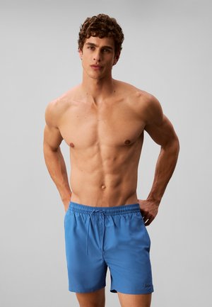 Hombre sin camiseta con cabello rizado que lleva pantalones cortos de baño azules, está de pie con las manos detrás de la espalda sobre un fondo liso.