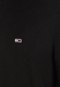 Zwarte gebreide trui met een klein wit, rood en blauw logo aan de linkerkant van de borst, met een gestructureerde stoffen oppervlakte.