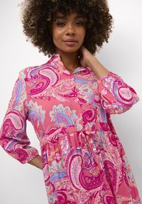 Roosa paisley kleit, millel on krae, ees nupud ja kokku keeratud vöökoht. Kangas on erksate värvide ja keerukate mustritega.