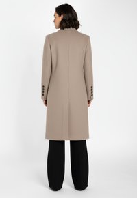 Beige lång jacka med en strukturerad design, texturerat tyg, snibbkrage, ryggslits och svarta knapppåsättningar på ärmarna, bärs över svarta byxor.