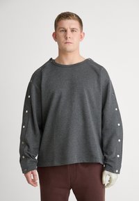 Grå langermet hettegenser med rund hals, trykknappdetaljer langs ermene og en avslappet passform. Stoffet ser mykt og teksturert ut.