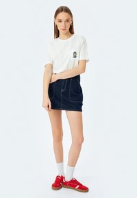 Witte katoenen t-shirt met een grafische print op de zak, gecombineerd met een donkerdenim mini-rok en rode sneakers met witte accenten.