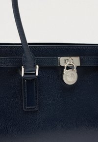 Tengerészkék bőr kézitáska texturált felülettel, ezüst színű fémalkatrészekkel, jellegzetes medállal és hosszú fogantyúkkal ellátott megformált alakzattal.