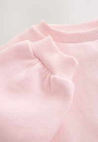Detailní záběr na světle růžovou mikinu z jemné látky, zobrazující žebrované manžety a výstřih s jemnou texturou a detaily švů.