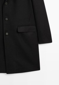 Cappotto di lana nera con un design sartoriale, parte anteriore con uno strappo singolo e sei bottoni, e una tasca laterale a sinistra. Texture liscia.