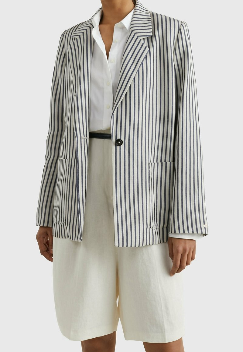 Femme portant une blouse blanche, un short blanc et un blazer à un seul bouton avec des rayures verticales bleu marine et blanc.