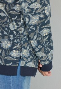 Persona vistiendo una blusa de manga larga de patrón floral azul con puños y dobladillo acanalados, combinada con pantalones vaqueros de denim azul claro.