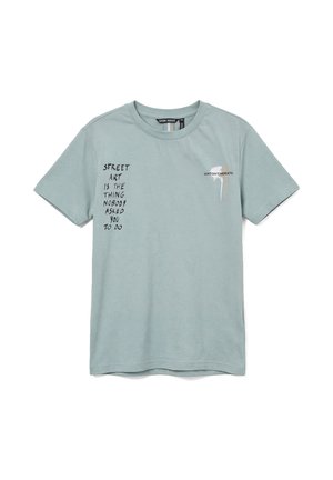T-shirt bleu clair à manches courtes, texte noir sur la poitrine gauche indiquant « Street art est la chose que personne ne vous a demandé de faire », avec un petit logo de taches de peinture.