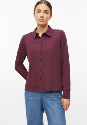 Frau trägt ein langärmliges, bordeauxrotes Hemd mit Knöpfen und blaue Jeans und steht vor einem weißen Hintergrund.
