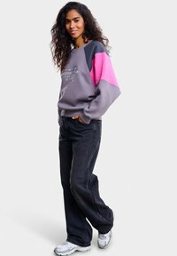 Šedá mikina s růžovými a tmavě modrými akcenty, s kulatým výstřihem a grafickým nápisem. Kombinována s tmavými džínami a stříbrnými teniskami.